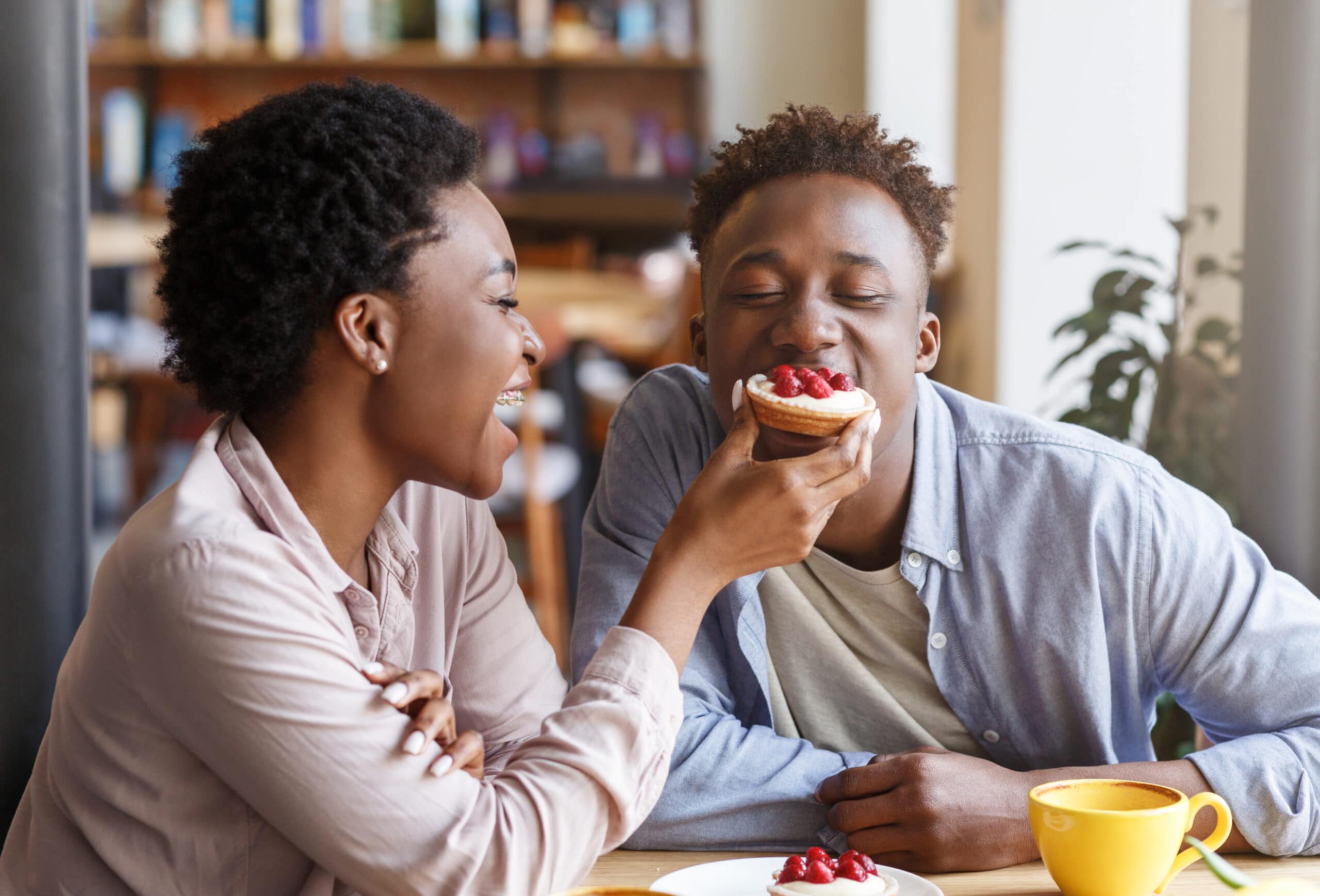African American girl feeding delicious sweet tartlet to her boyfriend at modern coffee shop
