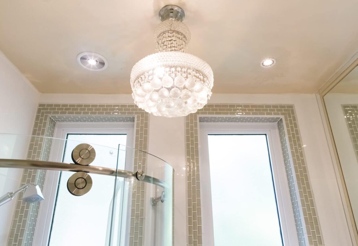 white luxe chandelier in bright white bathroom with green tile surrounding two rectangular windows and top of glass curved shower
