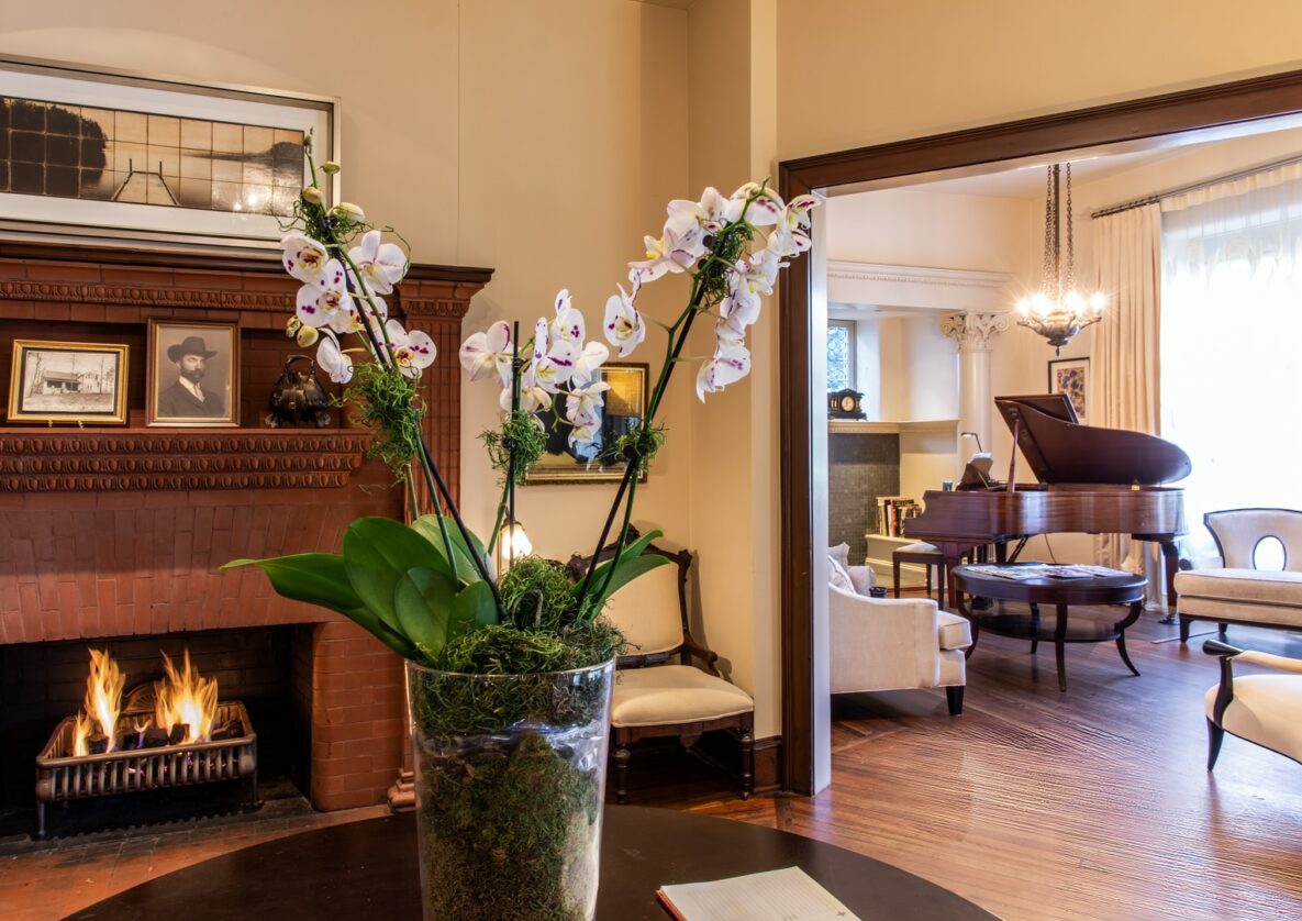 White orchids in glass vase on table in front of fire place and living room with white couches and brown piano