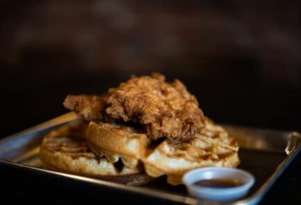 chicken and waffles on dark plate with dark background and syrup cup in foreground